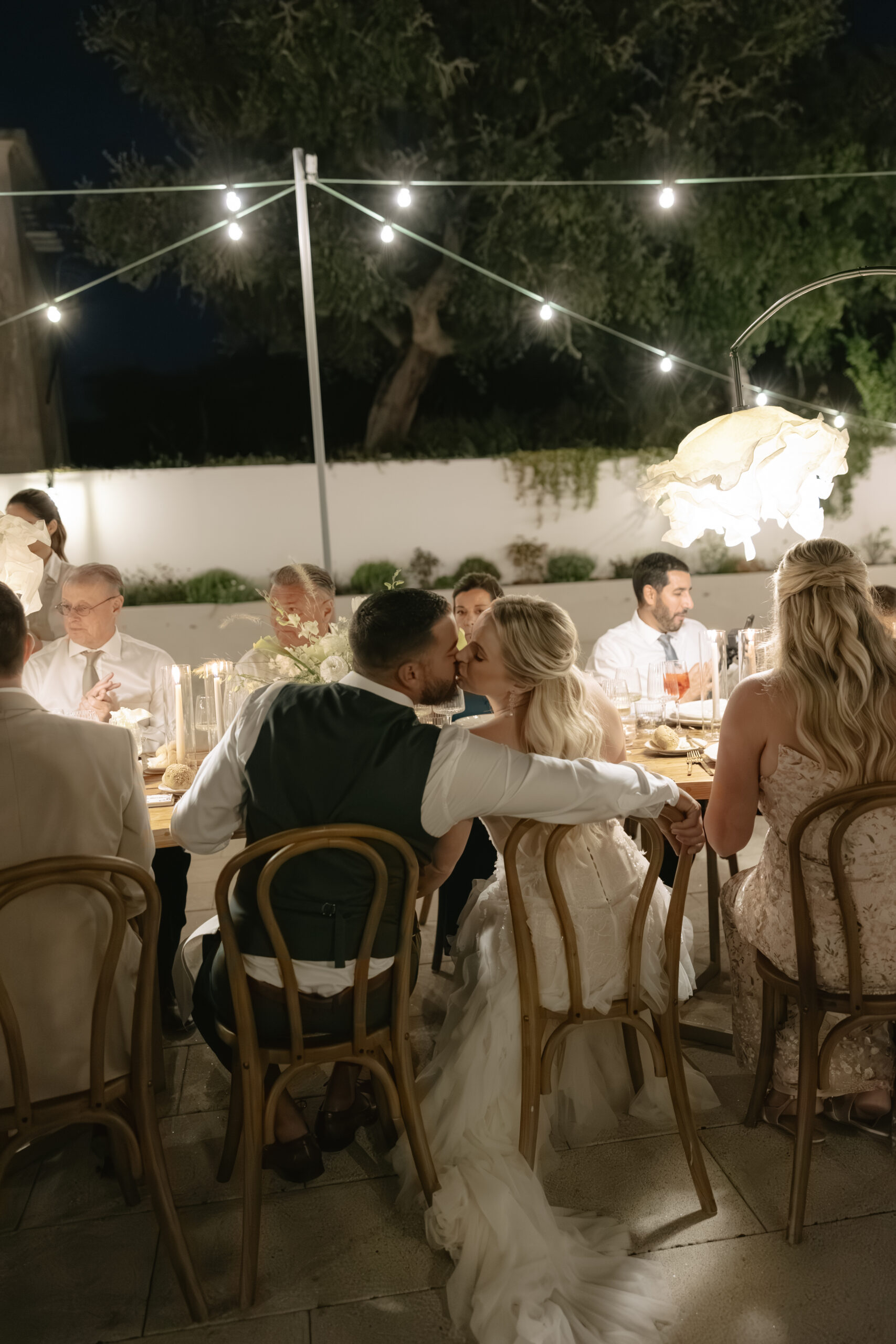 Bride and groom kissing at their wedding reception table under string lights
