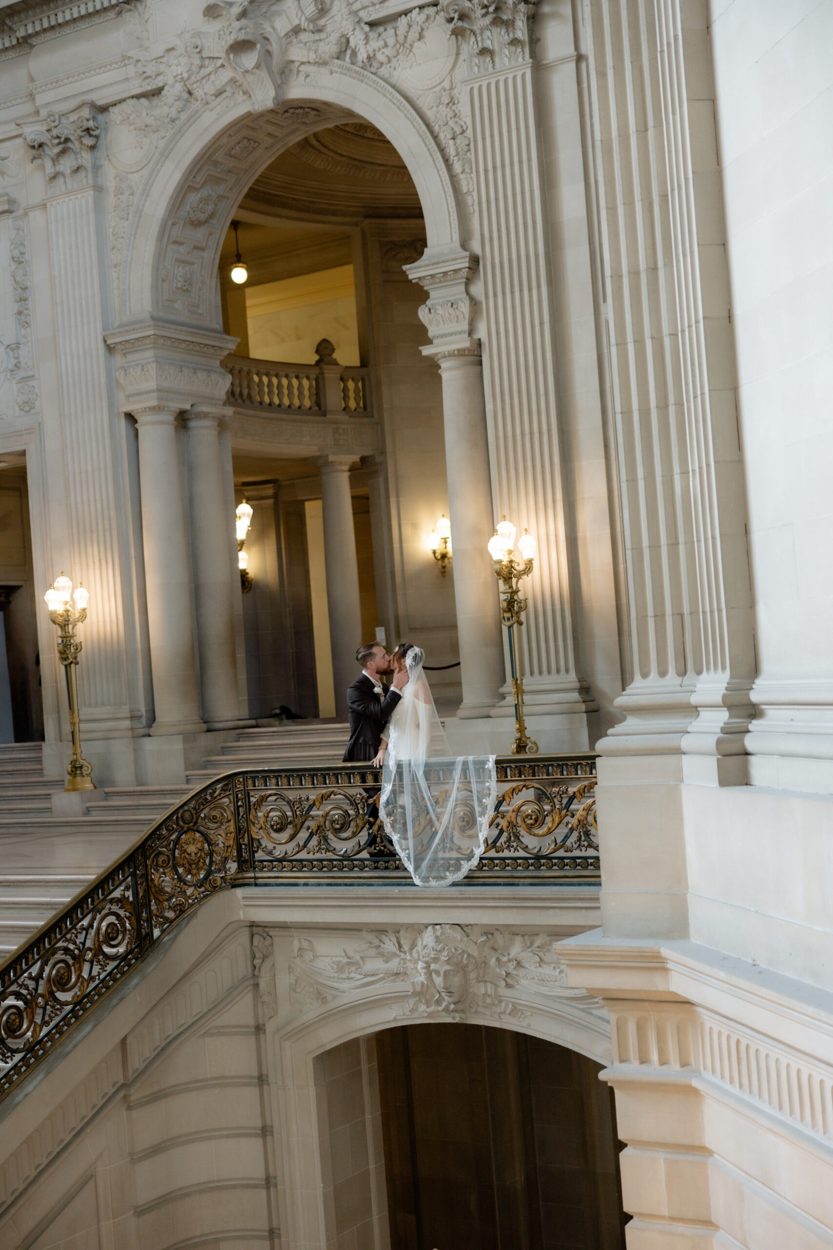 An editorial wedding photo of a bride and groom at San Francisco City Hall with the bride's veil draped over a railing