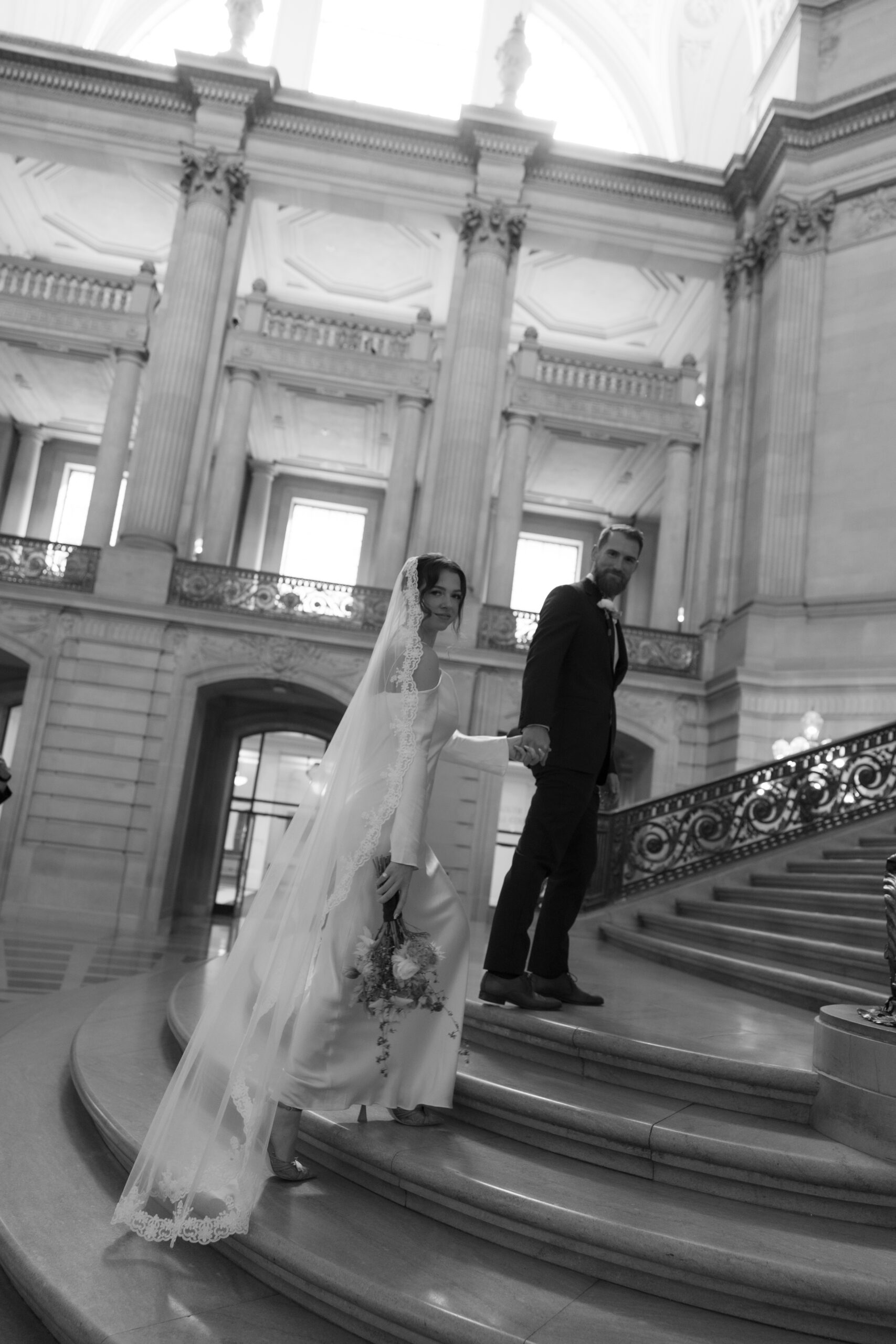 A black and white wedding photo of a bride and groom walking up the stairs at their San Francisco City Hall wedding