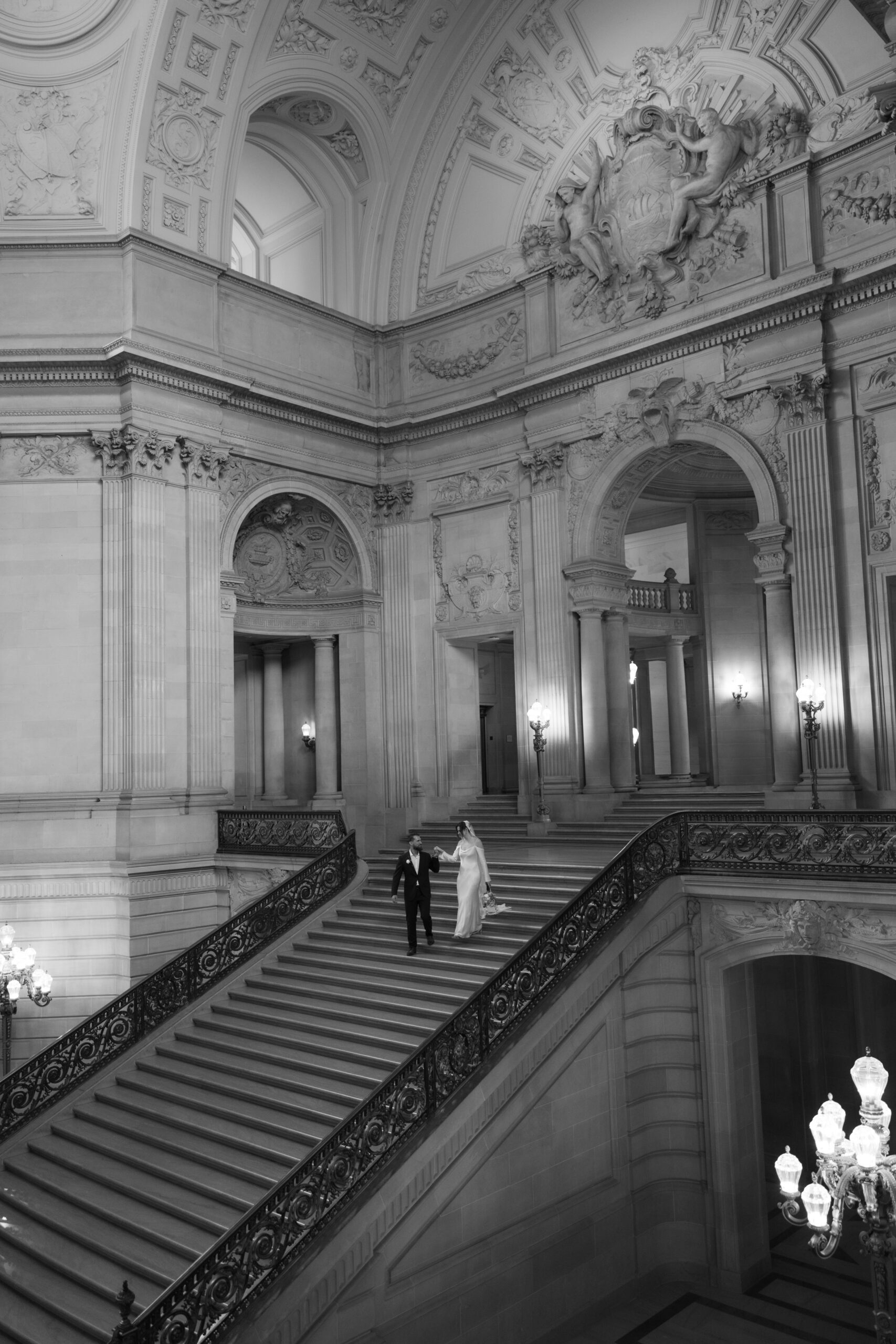A black and white photo of a bride and groom walking down a large staircase at their San Francisco City Hall wedding