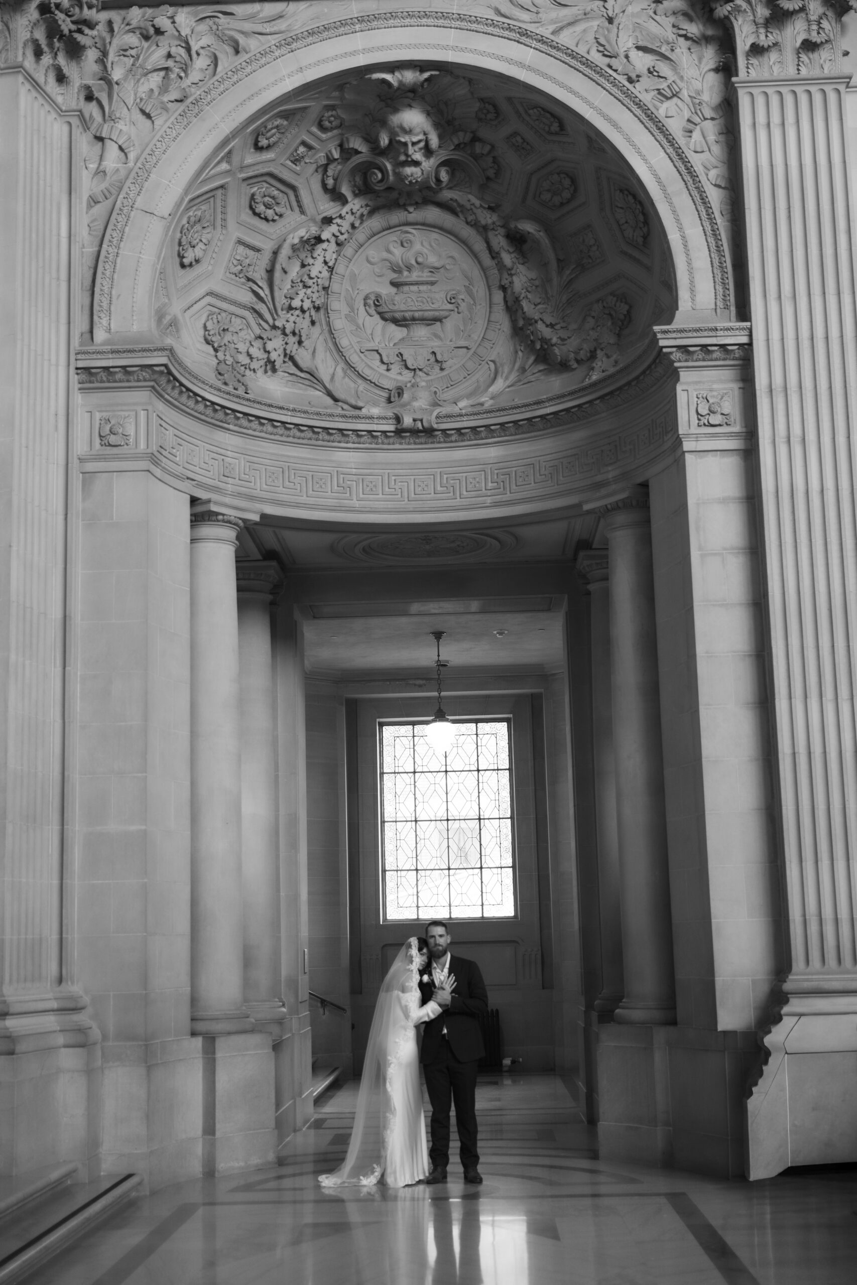 A black and white photo of a bride and groom standing in a hallway in San Francisco City Hall