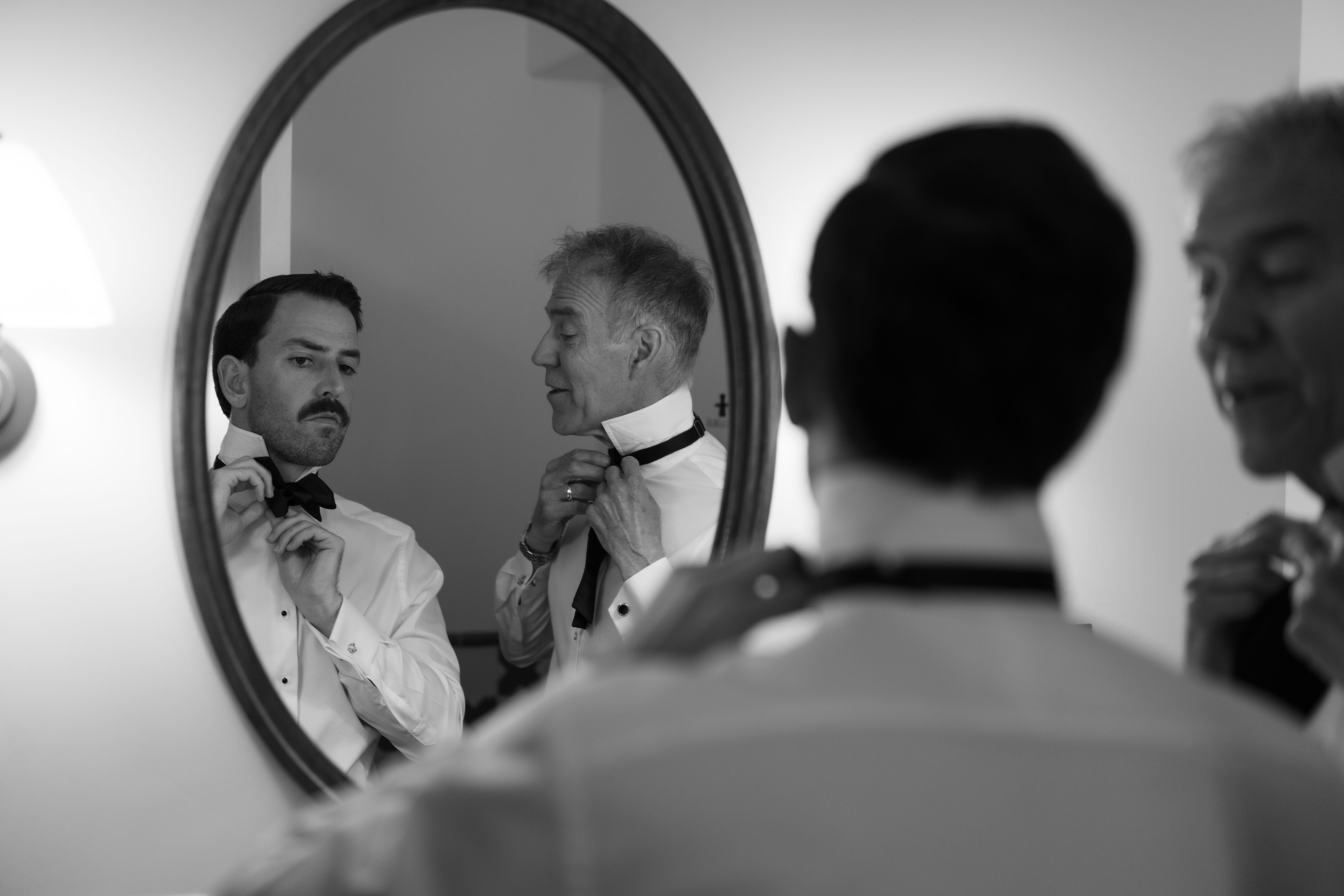 Photo of groom in mirror getting ready for wedding putting on his bowtie with his dad