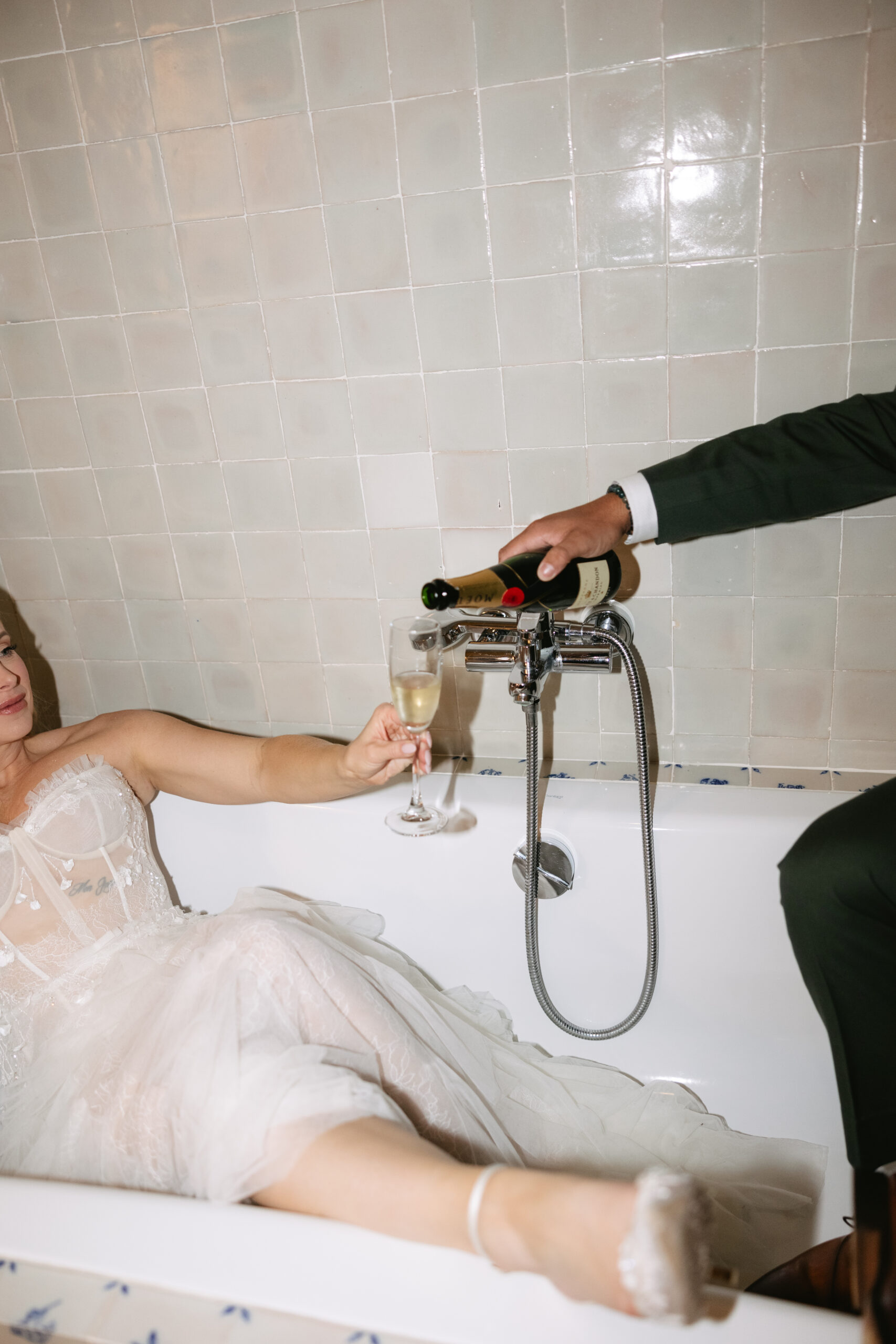 Editorial wedding photo of bride sitting in bathtub while groom pours champagne
