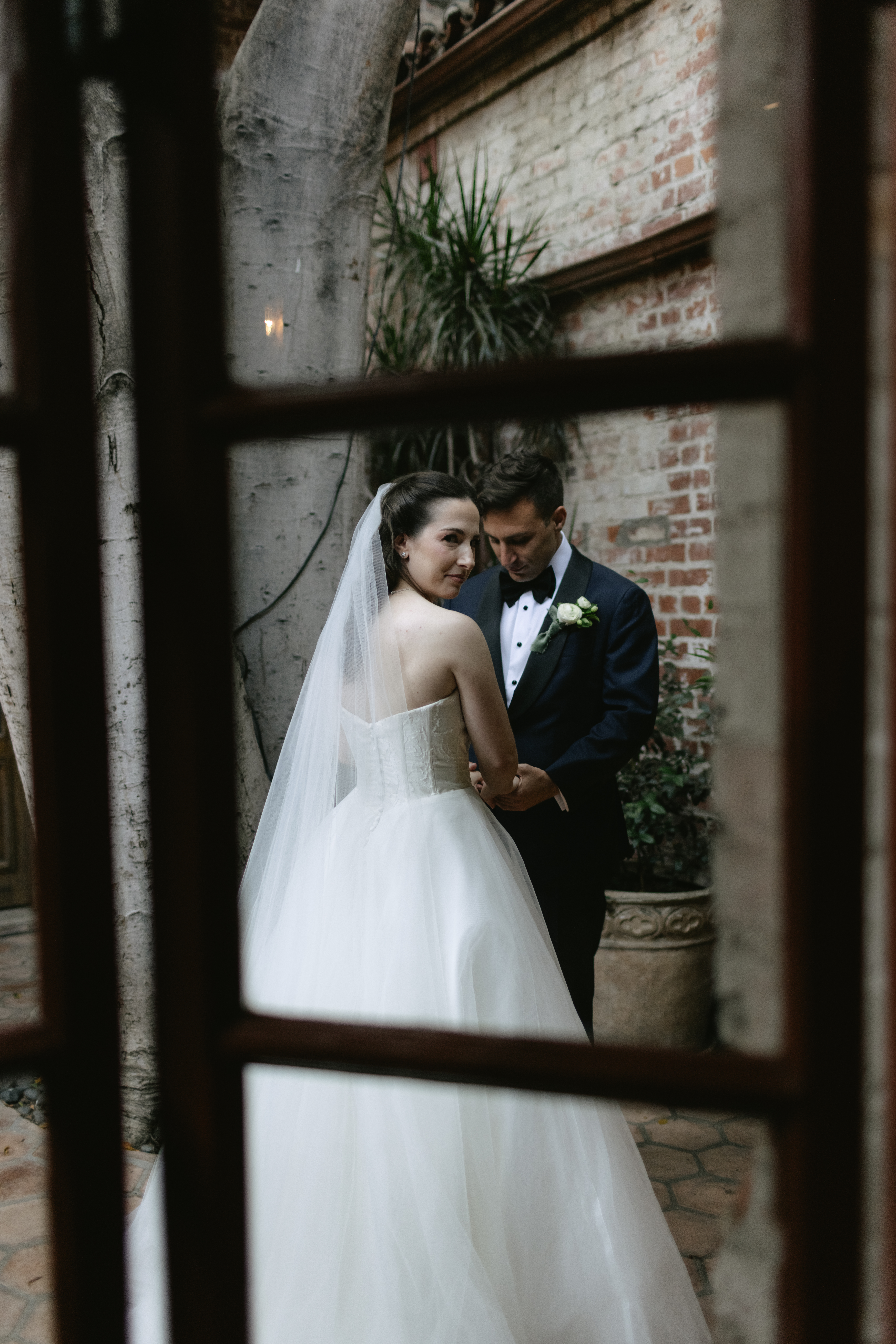 A bride and groom holding hands during a first look, captured through a window with documentary style wedding photography